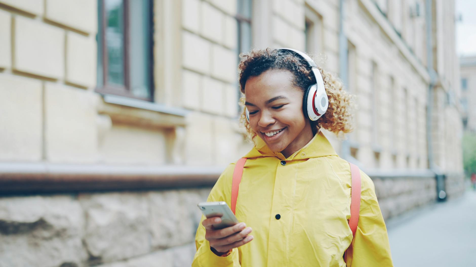 Uśmiechnięta kobieta w żółtym płaszczu słucha audiobooka w słuchawkach